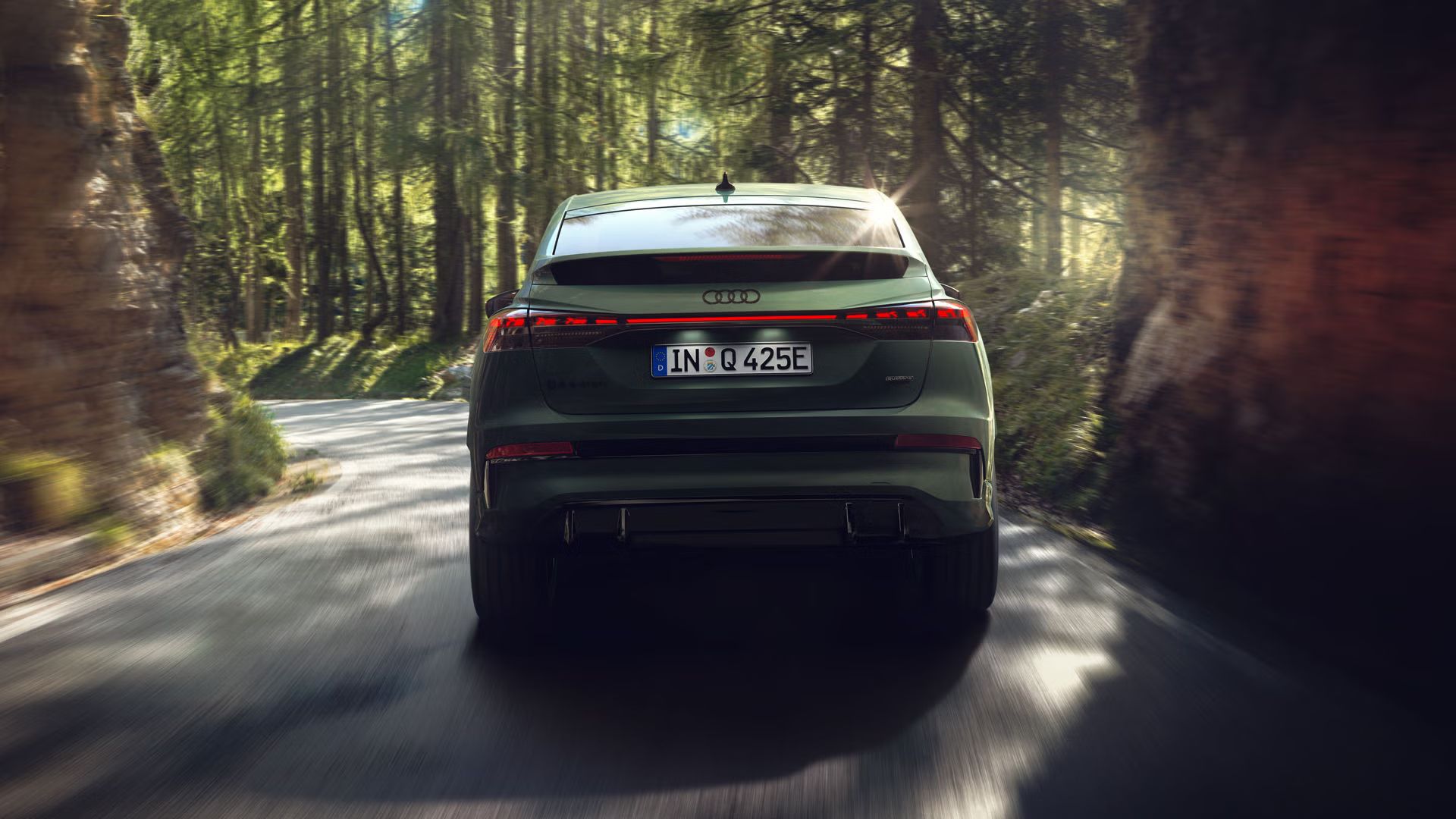 Der Q4 Sportback e-tron von hinten bei der Fahrt durch ein Waldstück, links und rechts der Straße ragen Felsen auf.