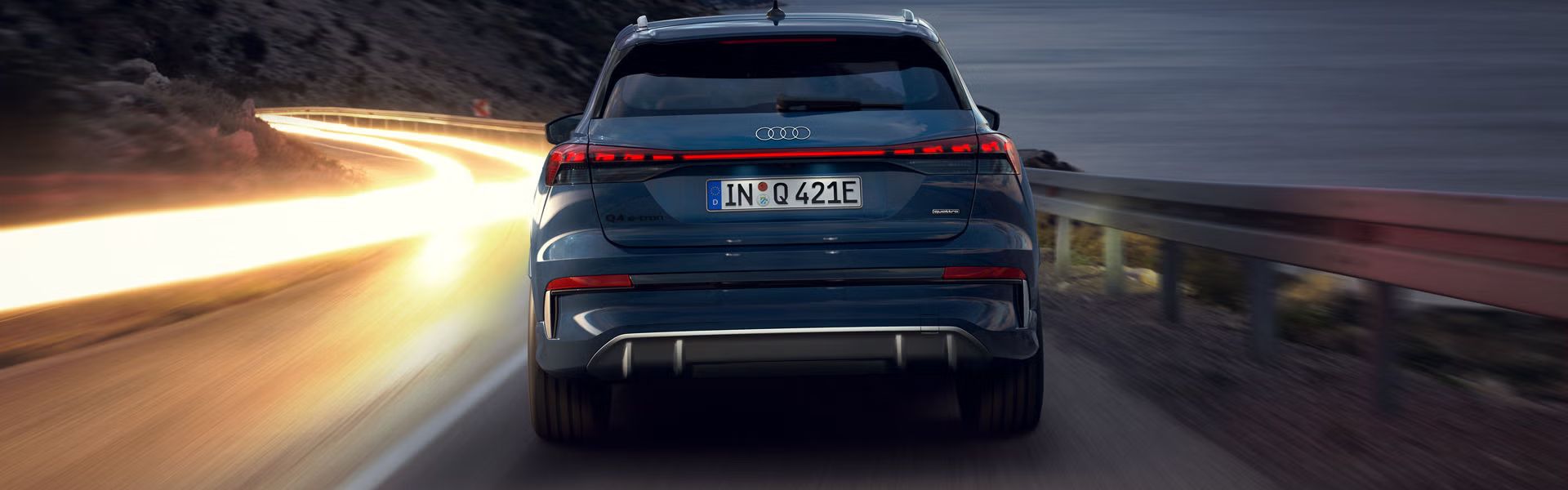 Der Q4 SUV e-tron von hinten bei Nacht auf einer abschüssigen Gebirgstraße, zur rechten das Meer. Auf der Gegenfahrbahn sind Lichtschleier entgegenkommender Fahrzeuge zu sehen. 