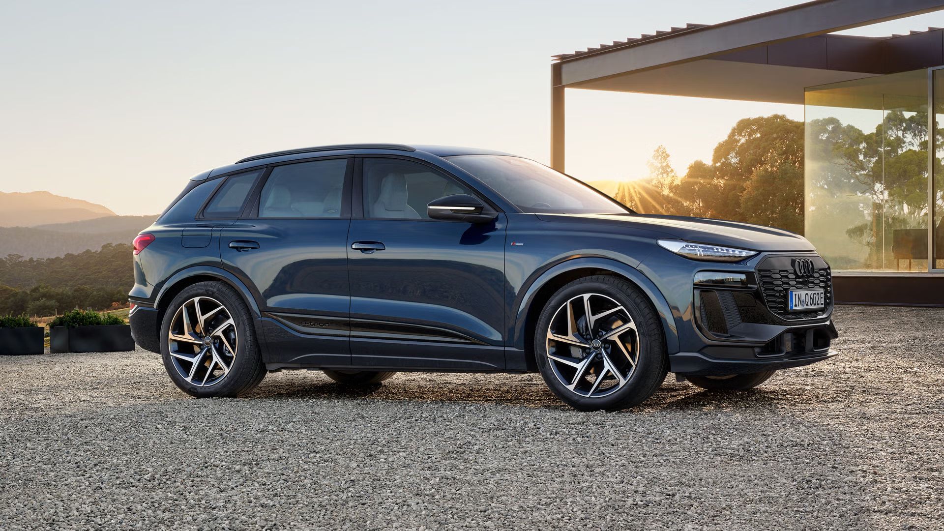 Der Q6 SUV e-tron steht seitlich auf der Kiesfläche vor einem modernen Haus und wird dabei von der untergehenden Sonne angestrahlt.