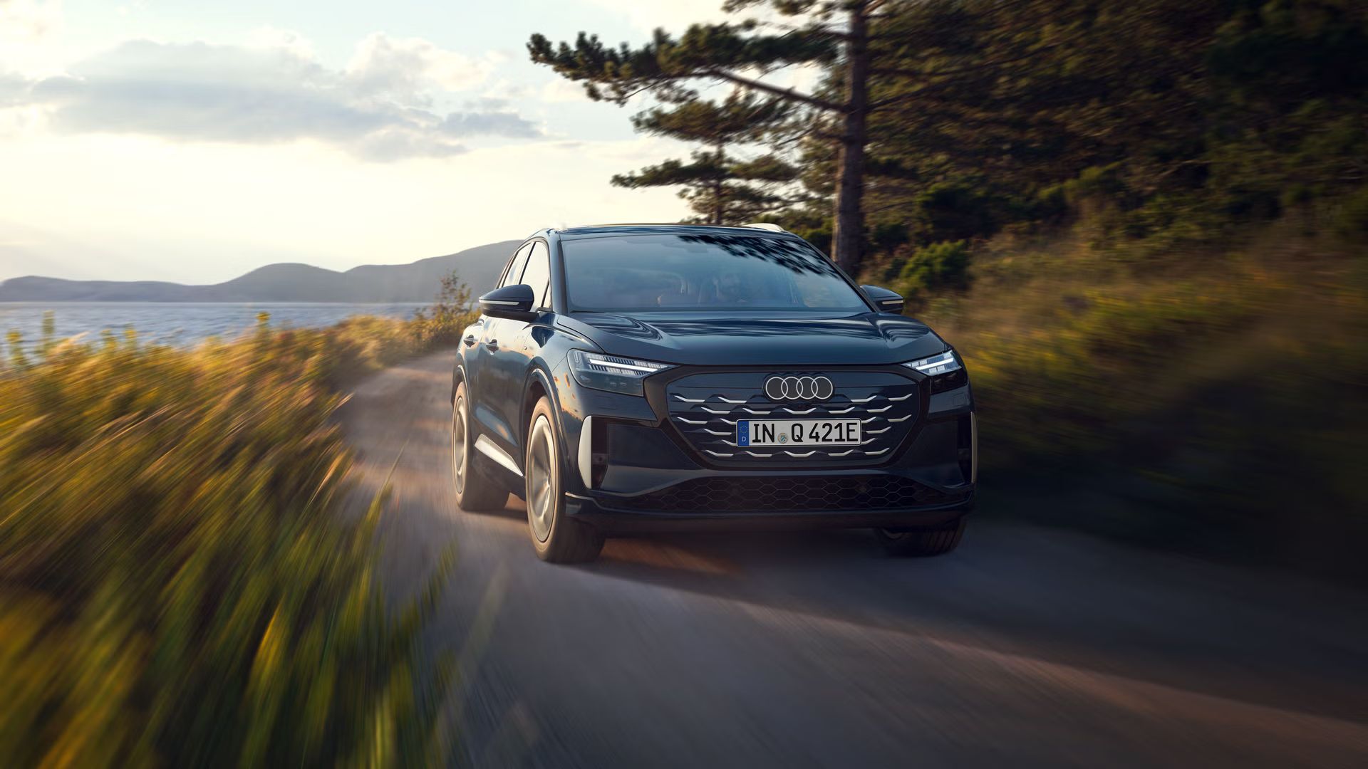 Der Q4 SUV e-tron schräg von vorn in voller Fahrt auf einer Landstraße an einem Waldsee, im Hintergrund eine bergige Landschaft.